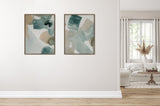 Two abstract framed canvases with green, blue, and beige brushstrokes hang side by side on a white wall in a bright living room with a cream sofa and wooden round table.
