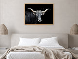 Framed canvas of a longhorn bull with curved cream horns and dark fur on a black background hanging above a modern bed with neutral beige bedding.